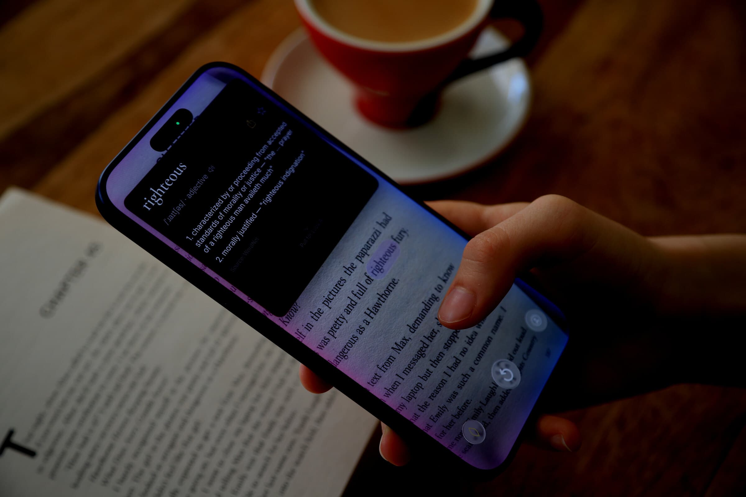 iPhone tilted in hand showing the Lupa live camera view and definition card over an open book