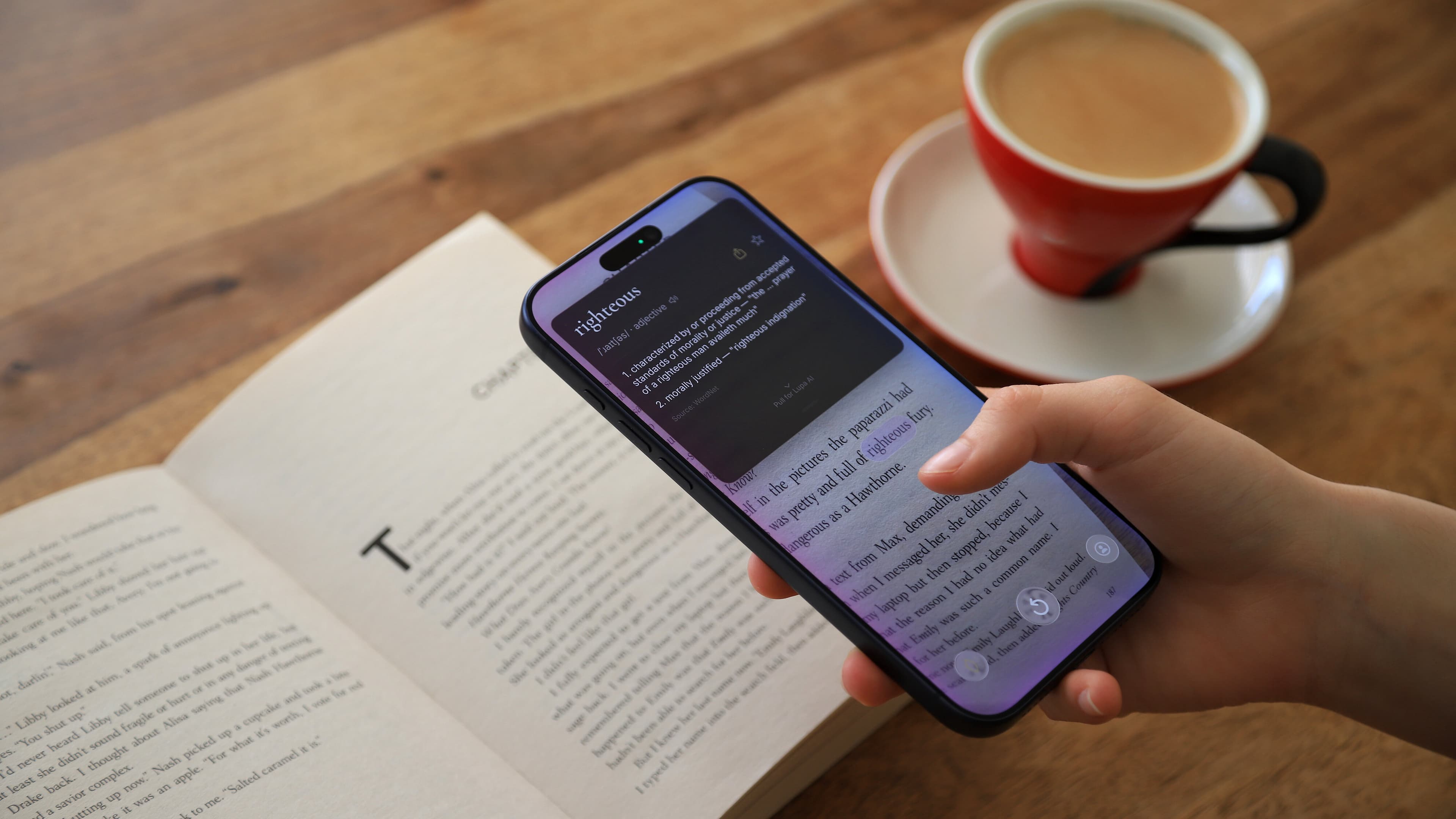 A reader holding an iPhone above an open novel, the Lupa app showing the definition of the word 'righteous', a red Lupa coffee cup beside the book